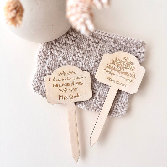 Two personalized wooden book mark bookends with engraved designs and text, displayed on a gray knitted blanket next to a ceramic vase with dried flowers.