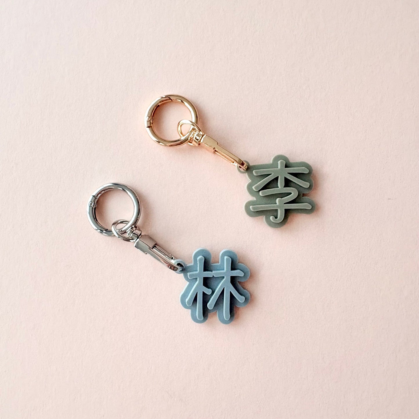 Two small keychain charms shaped like Chinese characters, one in light blue and one in muted green, attached to metal key rings on a pale pink background.