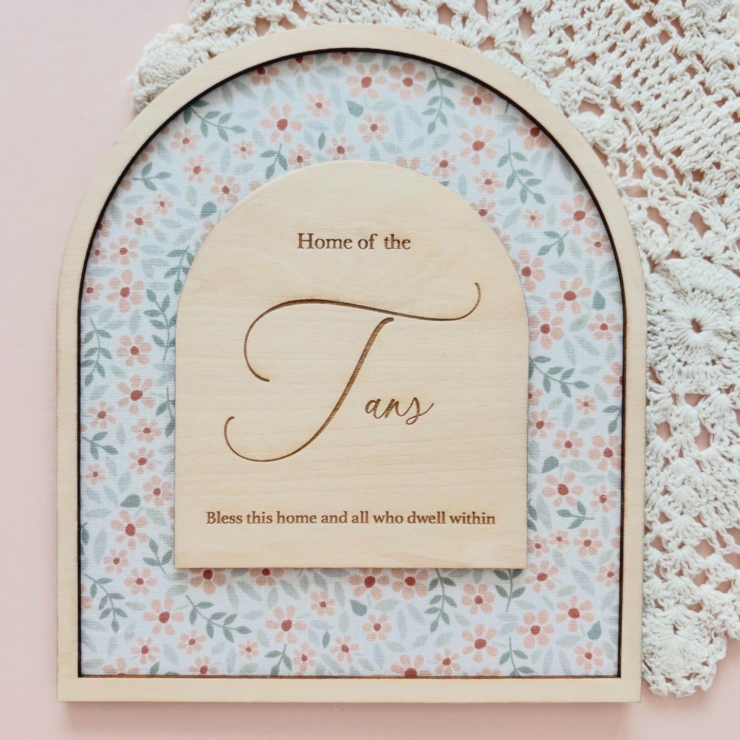 An arched wooden plaque with a floral patterned insert and a central wooden sign reading 'Home of the Fans' with additional decorative text, styled on a pink surface with a white lace doily in the background.