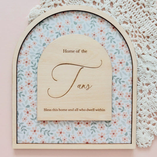 An arched wooden plaque with a floral patterned insert and a central wooden sign reading 'Home of the Fans' with additional decorative text, styled on a pink surface with a white lace doily in the background.