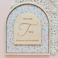 An arched wooden plaque with a floral patterned insert and a central wooden sign reading 'Home of the Fans' with additional decorative text, styled on a pink surface with a white lace doily in the background.