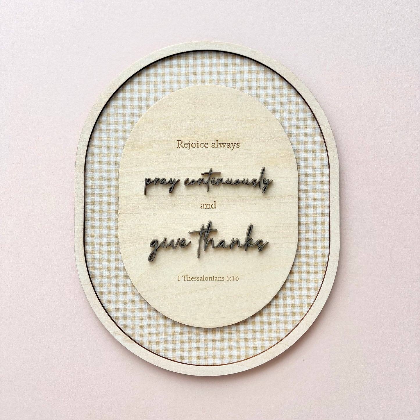 An oval wooden wall art piece with layered text reading 'Rejoice always, pray continually and give thanks' and '1 Thessalonians 5:16', set against a beige and white gingham patterned background, mounted on a light pink wall.