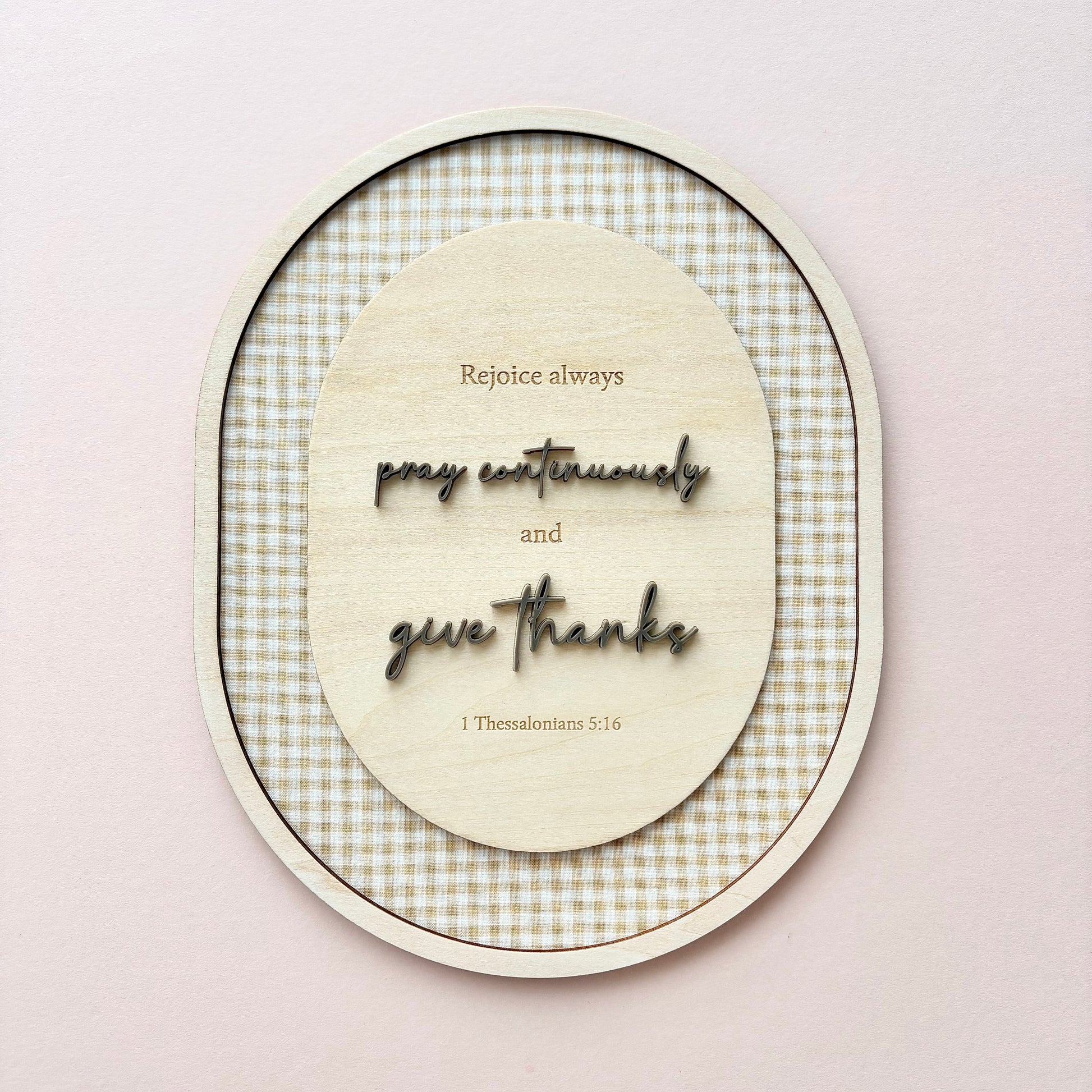 An oval wooden wall art piece with layered text reading 'Rejoice always, pray continually and give thanks' and '1 Thessalonians 5:16', set against a beige and white gingham patterned background, mounted on a light pink wall.