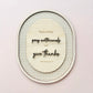 An oval wooden wall art piece with layered text reading 'Rejoice always, pray continually and give thanks' and '1 Thessalonians 5:16', set against a beige and white gingham patterned background, mounted on a light pink wall.
