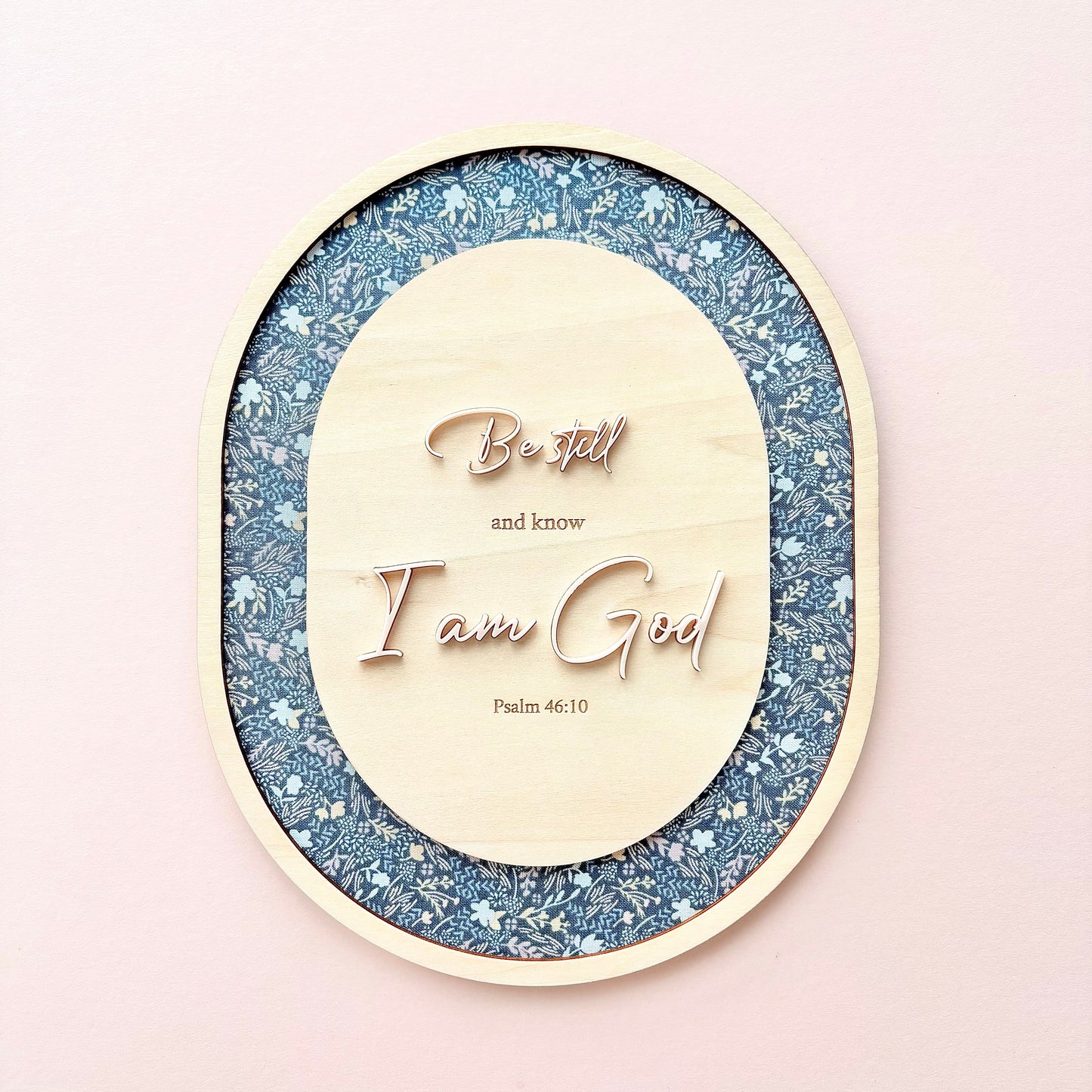 Oval wooden wall art featuring the carved text 'Best and know I am God Psalm 46:10' in rose-gold lettering, set against a glittery blue and white patterned background, displayed on a soft pink wall.