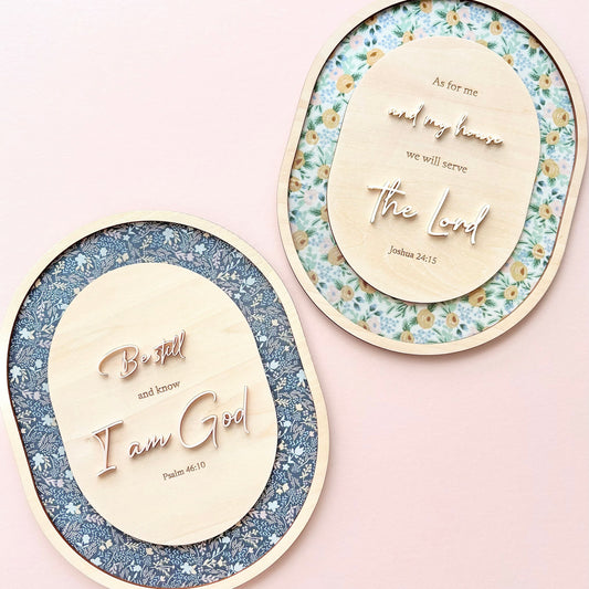 Two oval decorative wall art pieces with raised wooden text and floral-patterned backing, displayed on a soft pink background.