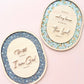 Two oval decorative wall art pieces with raised wooden text and floral-patterned backing, displayed on a soft pink background.