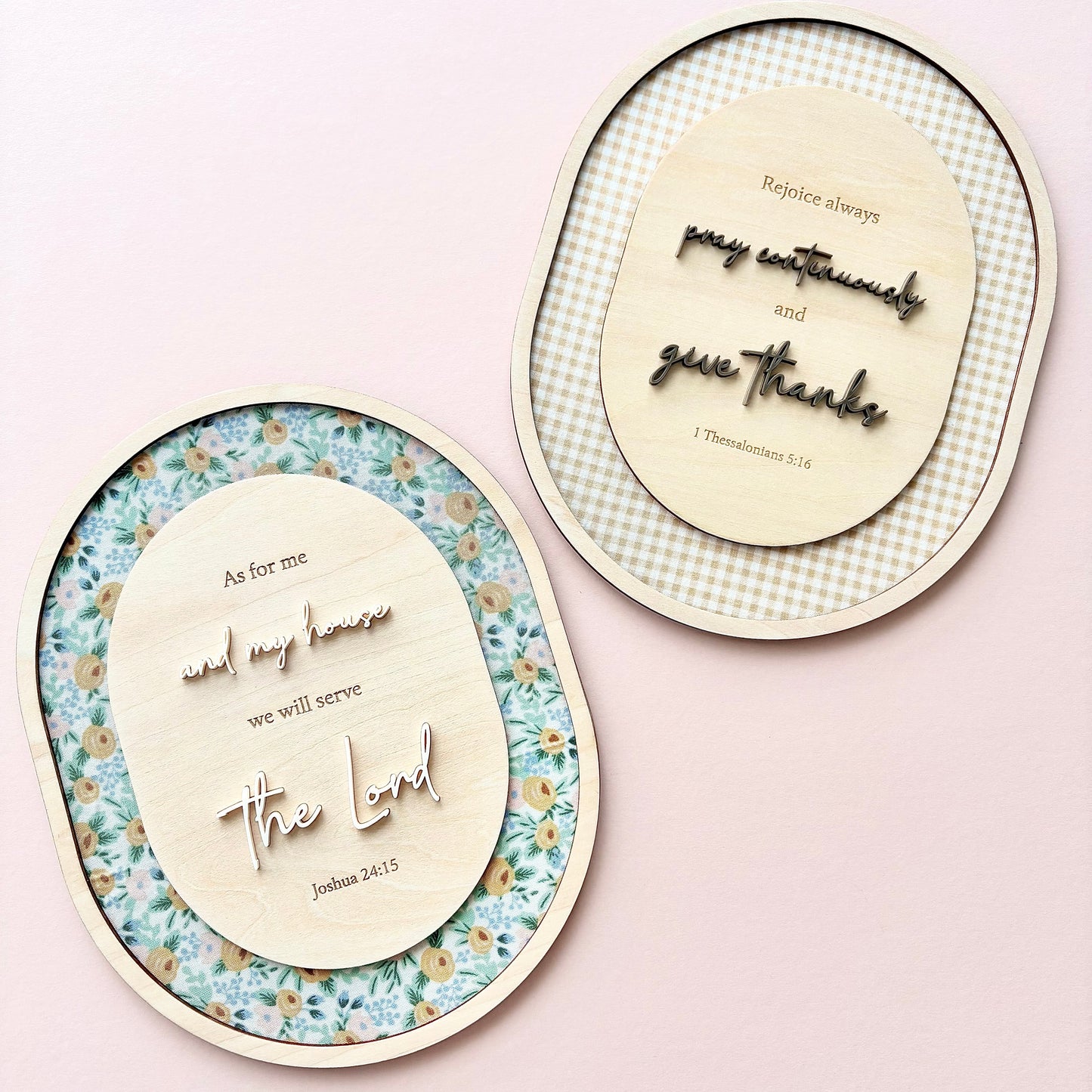Two oval decorative wall art pieces with wooden cutout lettering and floral or gingham borders, displayed on a soft pink background.