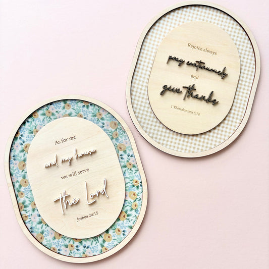 Two oval decorative wall art pieces with wooden cutout lettering and floral or gingham borders, displayed on a soft pink background.