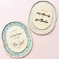 Two oval decorative wall art pieces with wooden cutout lettering and floral or gingham borders, displayed on a soft pink background.