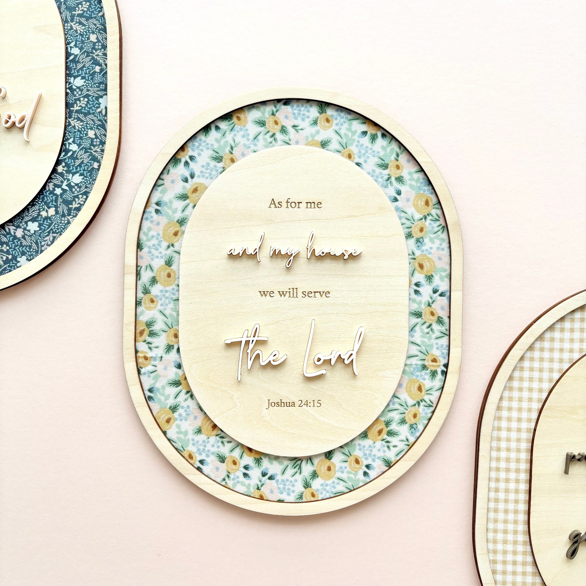Three oval wooden wall plaques with layered floral and gingham patterns, featuring engraved Bible verses including 'As for me and my house we will serve the Lord' and 'Joshua 24:15', displayed on a soft pink background.