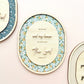Three oval wooden wall plaques with layered floral and gingham patterns, featuring engraved Bible verses including 'As for me and my house we will serve the Lord' and 'Joshua 24:15', displayed on a soft pink background.
