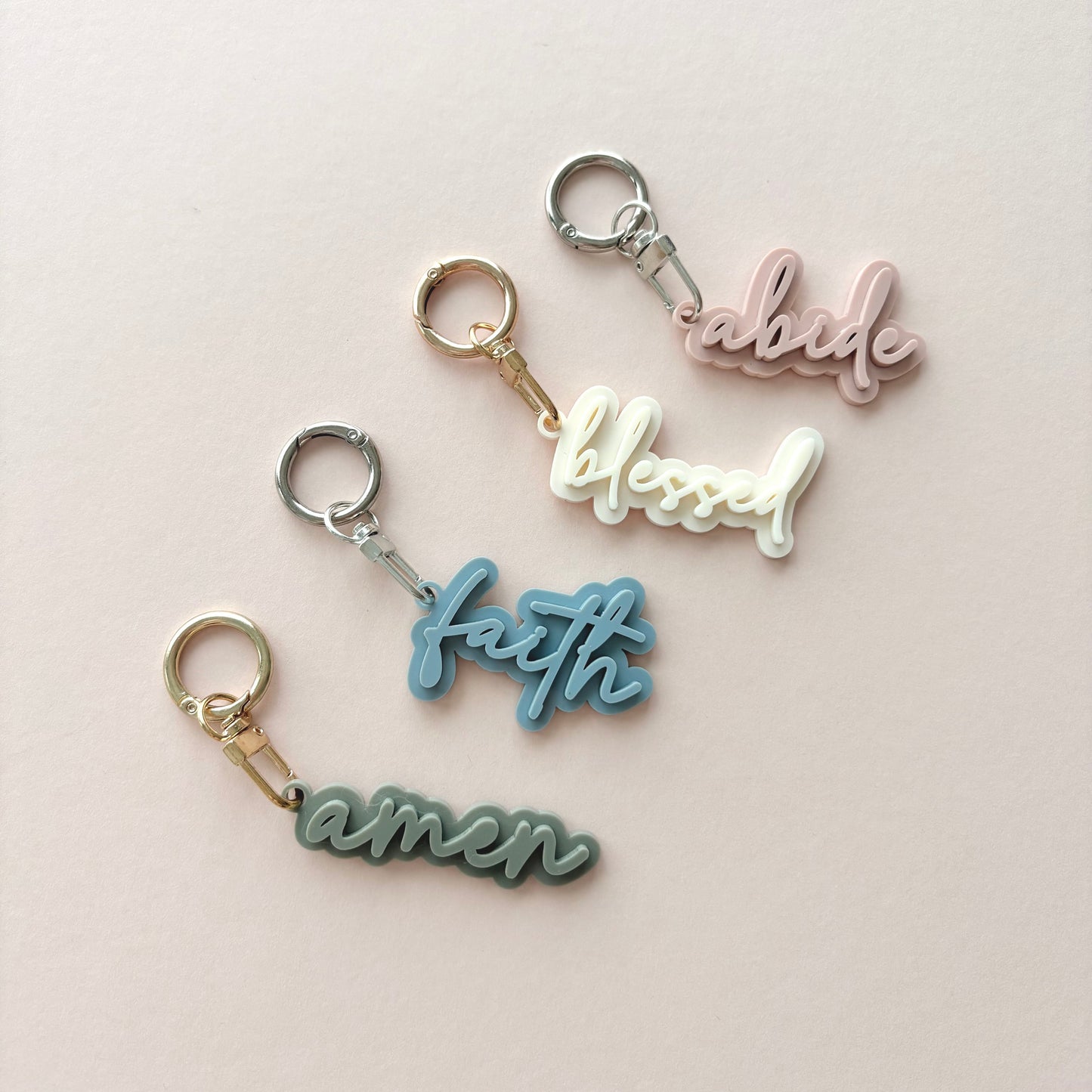 Four small script keychains on a pale pink background, each with a metal split ring and clasp in different colors: pink with gold, white with silver, light blue with gold, and muted green with silver. The words on the keychains read abide, blessed, faith, and amen.
