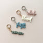 Four small script keychains on a pale pink background, each with a metal split ring and clasp in different colors: pink with gold, white with silver, light blue with gold, and muted green with silver. The words on the keychains read abide, blessed, faith, and amen.