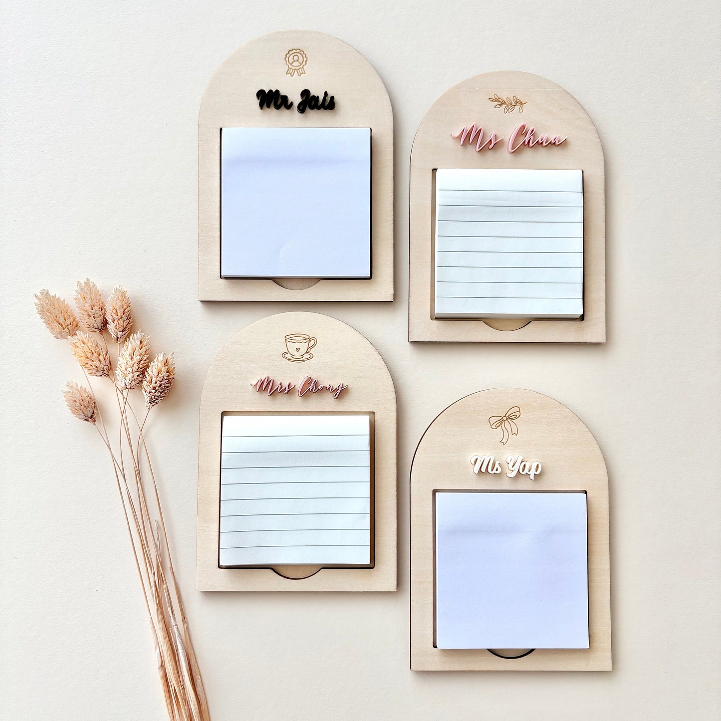 Four arched wooden notepad holders arranged in a 2x2 grid on a light beige background, with dried grass stems along the left side. The holders have engraved names or labels: Mr.
