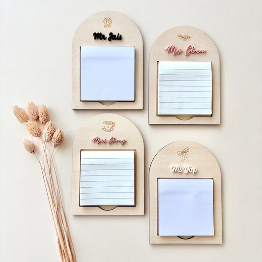 Four arched wooden notepad holders arranged in a 2x2 grid on a light beige background, with dried grass stems along the left side. The holders have engraved names or labels: Mr.