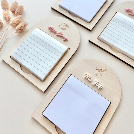 Four arched wooden notepad holders arranged on a light beige surface, each holding a white sticky note pad. Decorative dried stems appear on the left, and the notepads have small engraved or printed names including 'Miss Yap' and 'Ms.