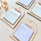 Four arched wooden notepad holders arranged on a light beige surface, each holding a white sticky note pad. Decorative dried stems appear on the left, and the notepads have small engraved or printed names including 'Miss Yap' and 'Ms.