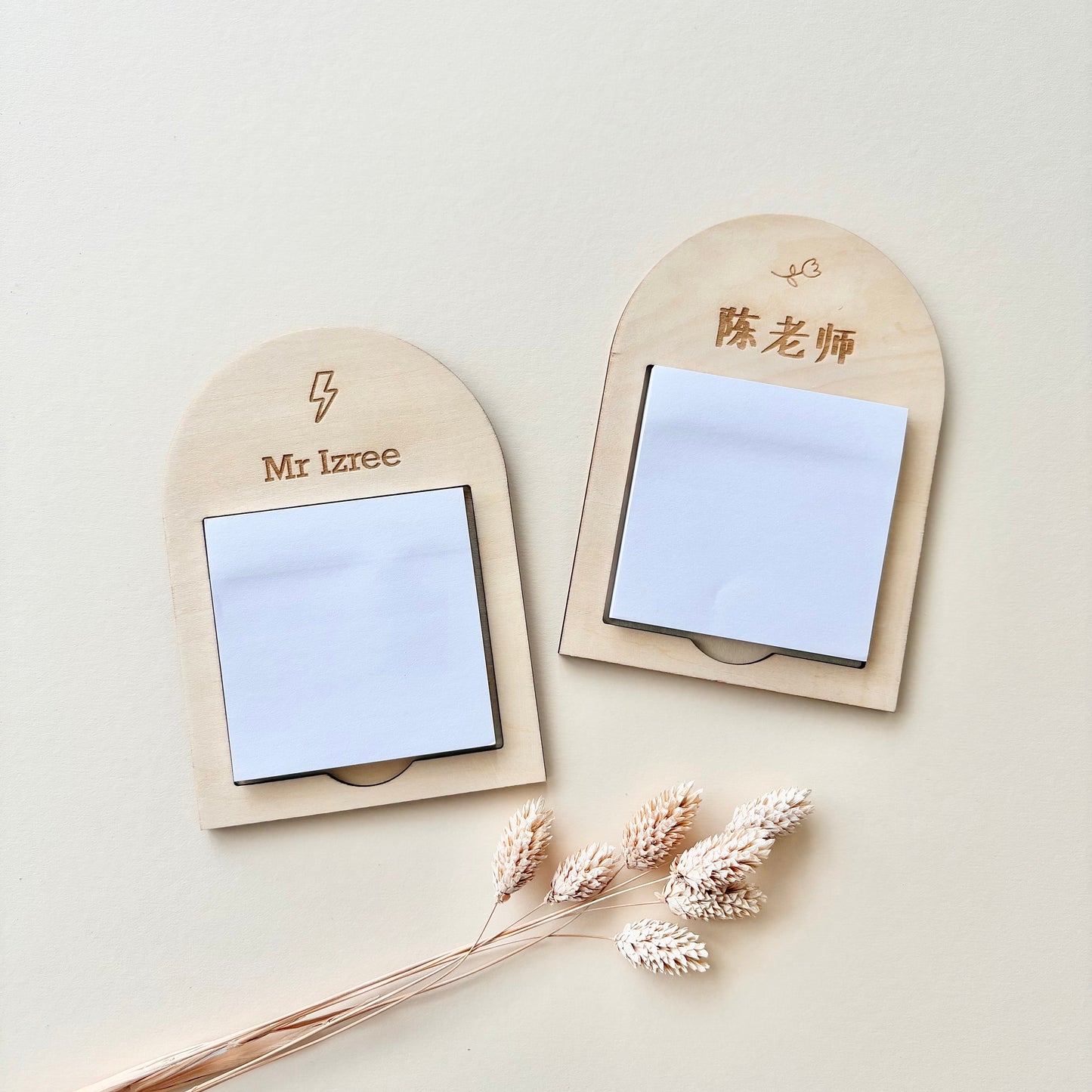 Two personalized wooden name badges with sticky note holders, displayed on a light beige background with dried decorative grass stems. One badge is engraved with 'Mr Izree' and a lightning bolt, and the other with '歕品团' (Teacher Chen) and a small heart.