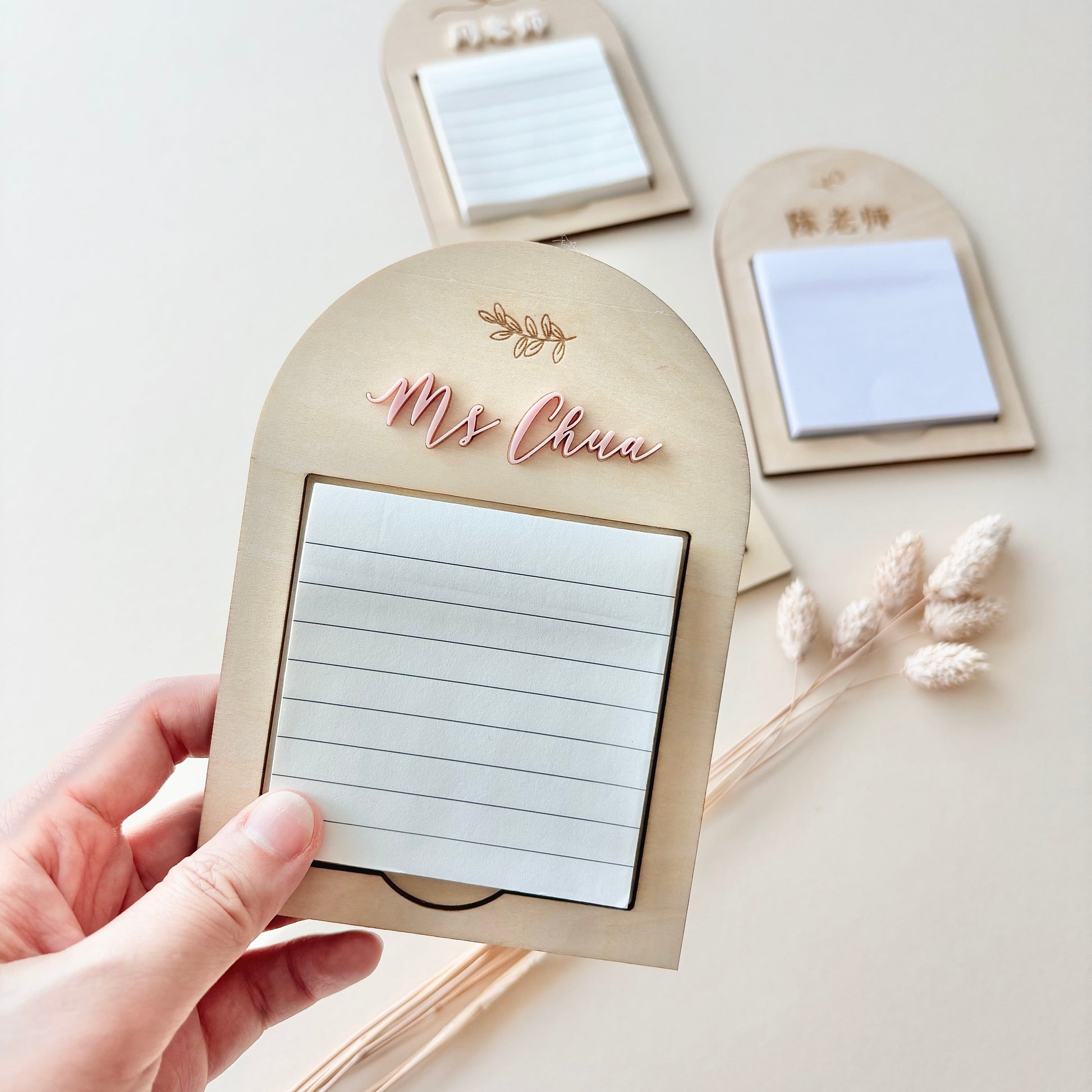 A hand holds a wooden arched notepad holder engraved with 'Mrs. Clara' in rose gold script, showing a white lined notepad inside.