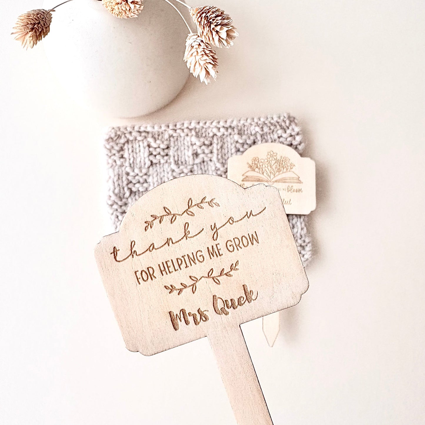 A hand holds a personalized wooden thank-you sign with the engraved text 'thank you FOR HELPING ME GROW Mrs Quek' and a small tree illustration. In the background, a knitted beige item and a small wooden tag rest against a light neutral surface, with a white ceramic vase and dried flowers above.