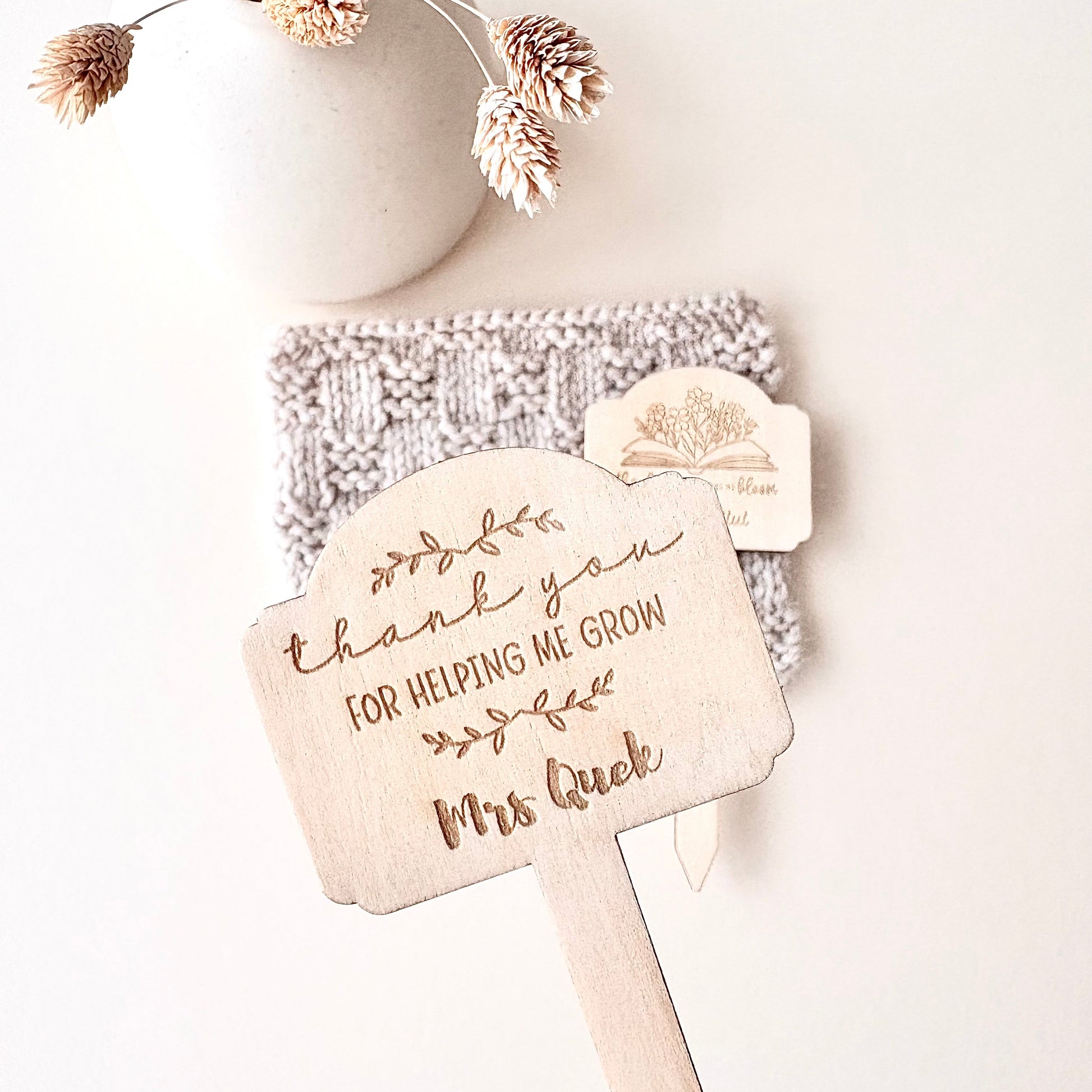 A hand holds a personalized wooden thank-you sign with the engraved text 'thank you FOR HELPING ME GROW Mrs Quek' and a small tree illustration. In the background, a knitted beige item and a small wooden tag rest against a light neutral surface, with a white ceramic vase and dried flowers above.