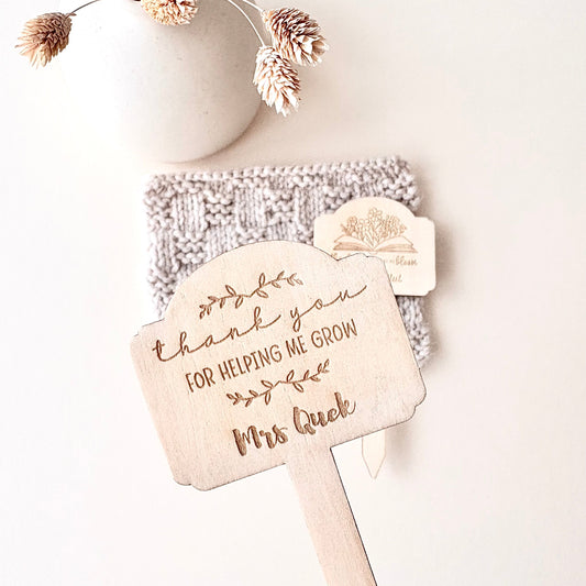 A hand holds a personalized wooden thank-you sign with the engraved text 'thank you FOR HELPING ME GROW Mrs Quek' and a small tree illustration. In the background, a knitted beige item and a small wooden tag rest against a light neutral surface, with a white ceramic vase and dried flowers above.