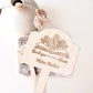 A close-up of a wooden thank-you sign on a stick with a personalized engraving of an open book and flowers, reading 'thank you for helping me bloom Mam Nailul'. The sign is held in a hand against a clean white background with a ceramic vase and dried flowers as styled props.
