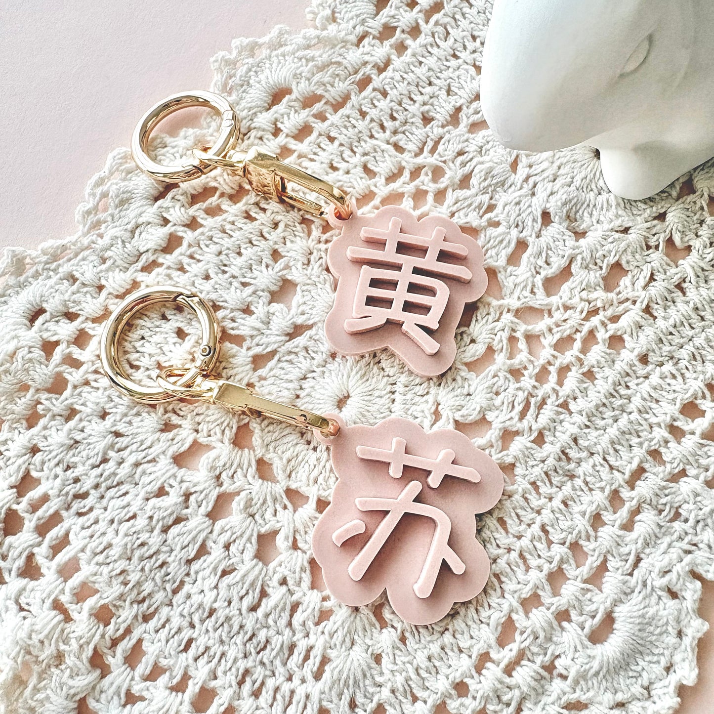 Two pastel pink flower-shaped keychains with gold-tone rings and clasps, each featuring a raised Chinese character, displayed on a white crocheted lace mat over a pale pink background.