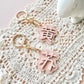 Two pastel pink flower-shaped keychains with gold-tone rings and clasps, each featuring a raised Chinese character, displayed on a white crocheted lace mat over a pale pink background.