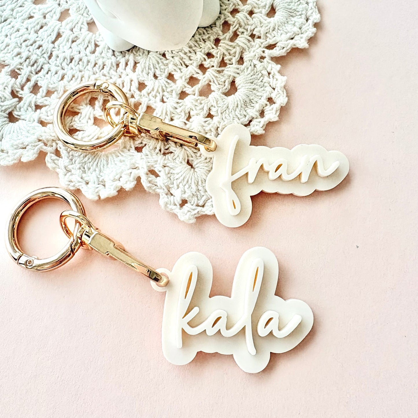 Two personalized acrylic keychains with gold-tone keyrings and clips, featuring the names 'frank' and 'kala' in white cursive script, styled on a pink background with a white lace doily.