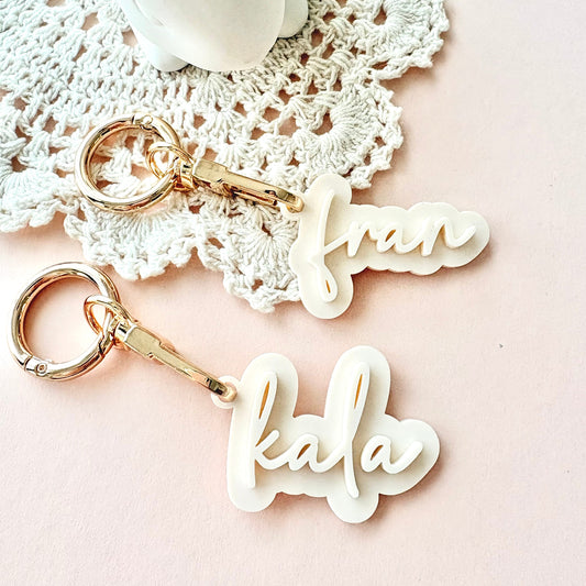 Two personalized acrylic keychains with gold-tone keyrings and clips, featuring the names 'frank' and 'kala' in white cursive script, styled on a pink background with a white lace doily.