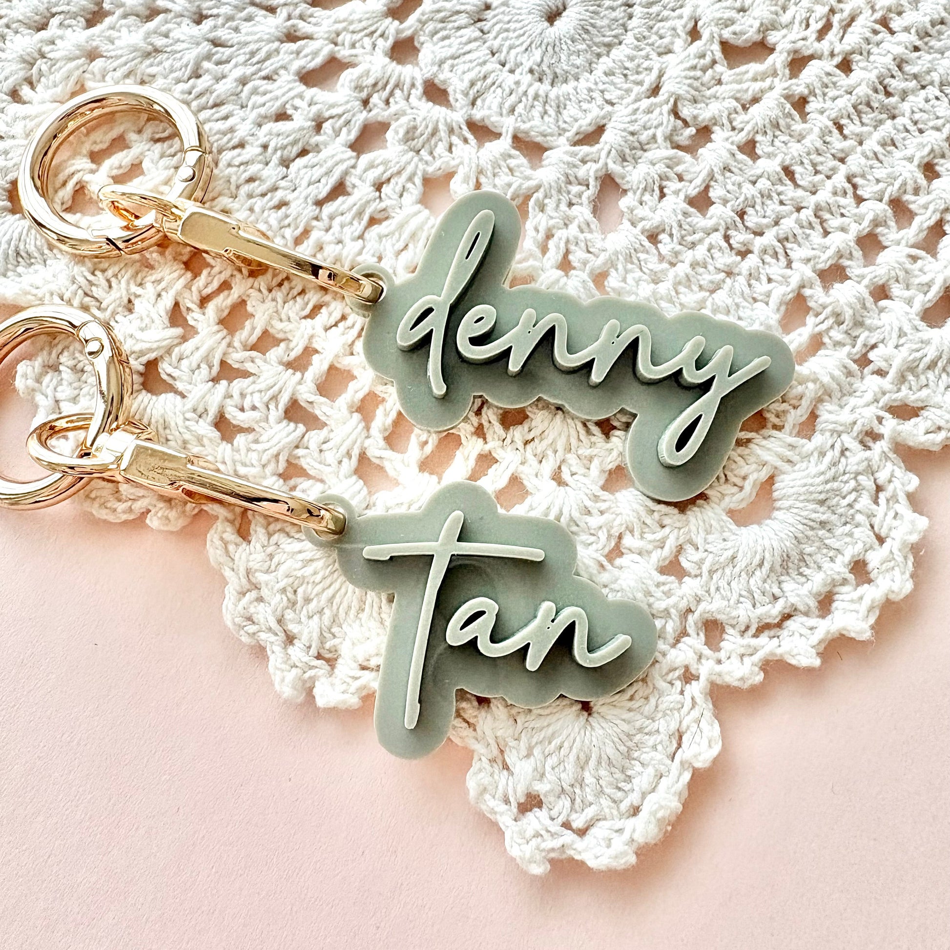 Two personalized keychains with gold-tone split rings and clips, featuring custom names 'denny' and 'tan' in white script on a sage green background, displayed on a white crocheted doily over a soft pink surface.