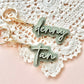 Two personalized keychains with gold-tone split rings and clips, featuring custom names 'denny' and 'tan' in white script on a sage green background, displayed on a white crocheted doily over a soft pink surface.