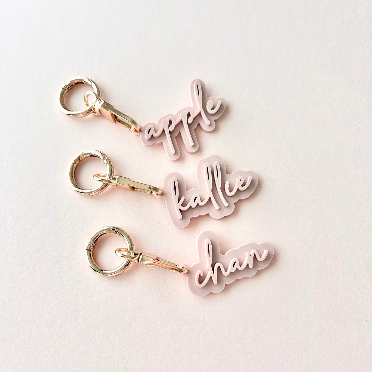 Three personalized acrylic keychains with gold-tone keyrings and clips, featuring the names 'apple', 'kallie', and 'chan' in light pink cursive script, arranged vertically on a light off-white background.