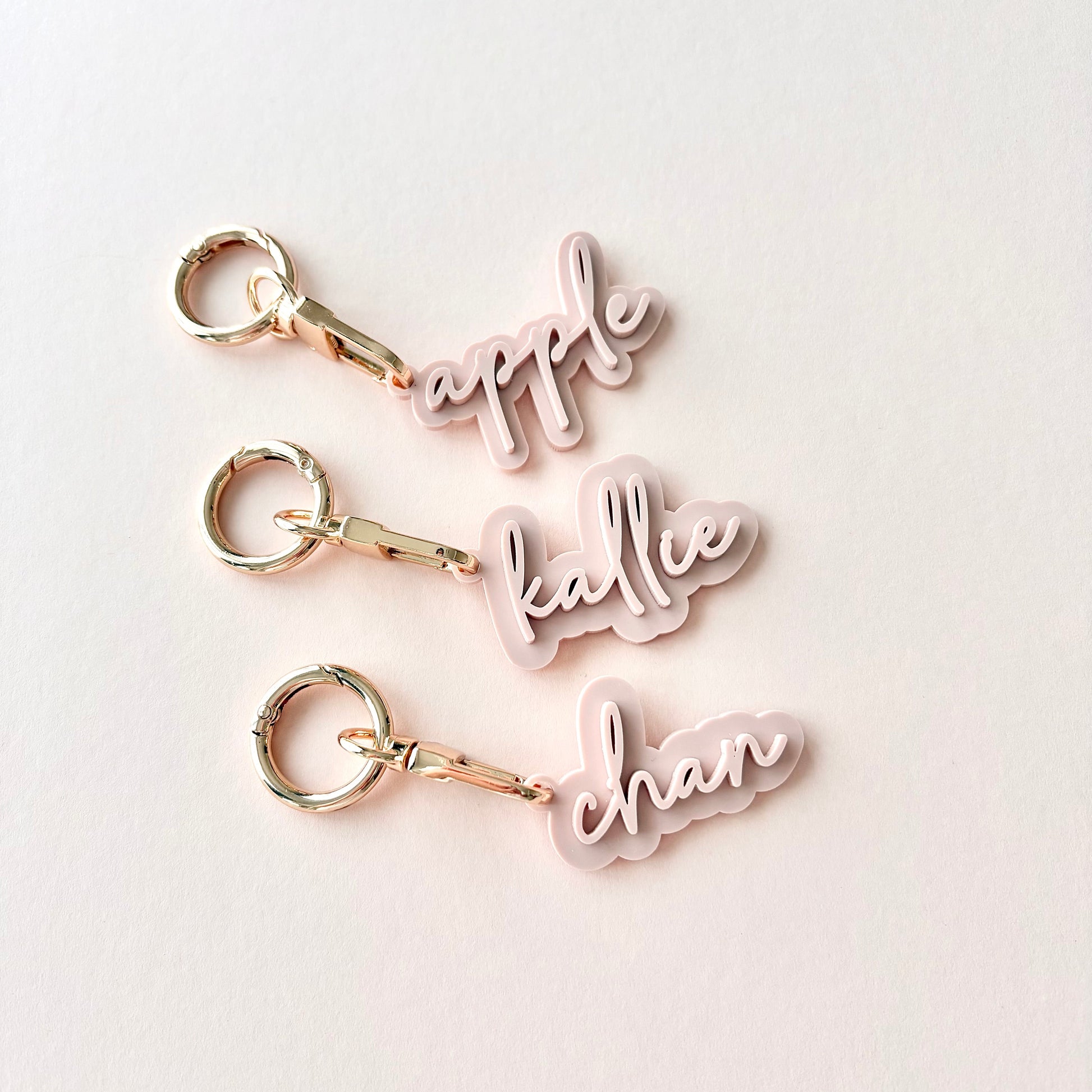 Three personalized acrylic keychains with gold-tone keyrings and clips, featuring the names 'apple', 'kallie', and 'chan' in light pink cursive script, arranged vertically on a light off-white background.