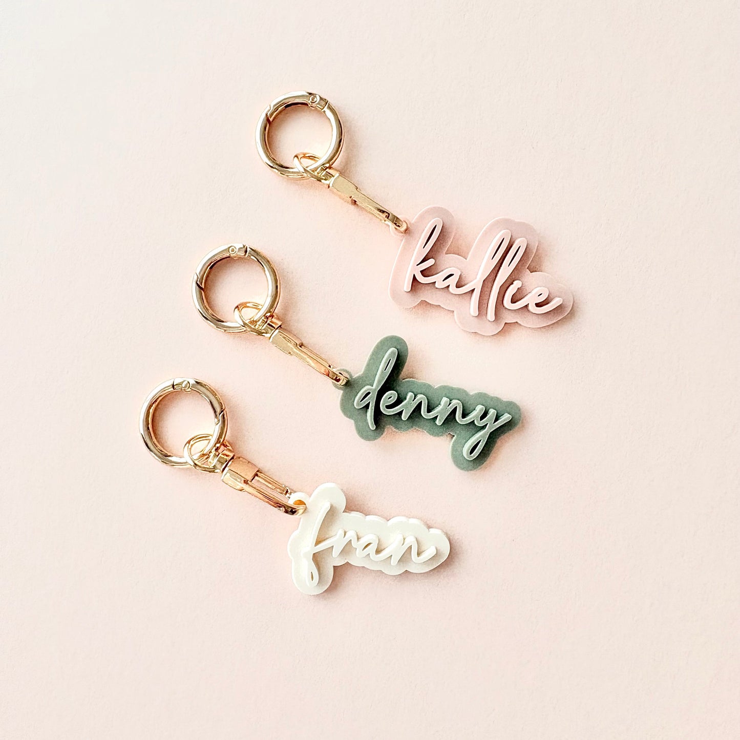 Three personalized acrylic keychains spelling 'kallie', 'denny', and 'frank' in pink, sage green, and white cursive fonts, each with a gold split ring and clasp, arranged on a light pink background.
