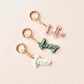 Three personalized acrylic keychains spelling 'kallie', 'denny', and 'frank' in pink, sage green, and white cursive fonts, each with a gold split ring and clasp, arranged on a light pink background.