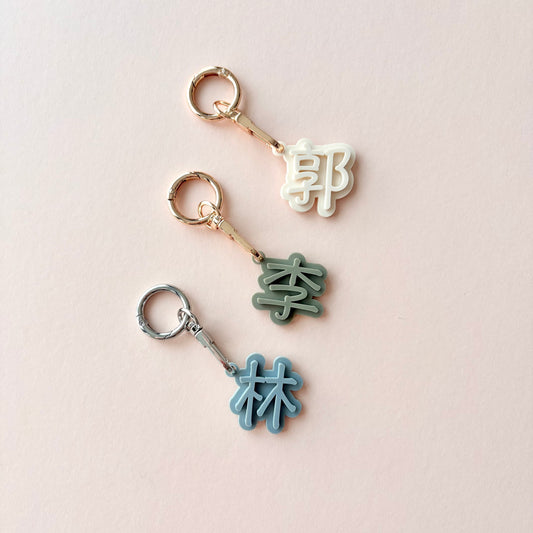 Three small keychain charms with Chinese characters arranged vertically on a pale pink background. The top charm is cream with a gold ring and connector, the middle is sage green with a gold connector, and the bottom is light blue with a silver connector.