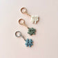 Three small keychain charms with Chinese characters arranged vertically on a pale pink background. The top charm is cream with a gold ring and connector, the middle is sage green with a gold connector, and the bottom is light blue with a silver connector.
