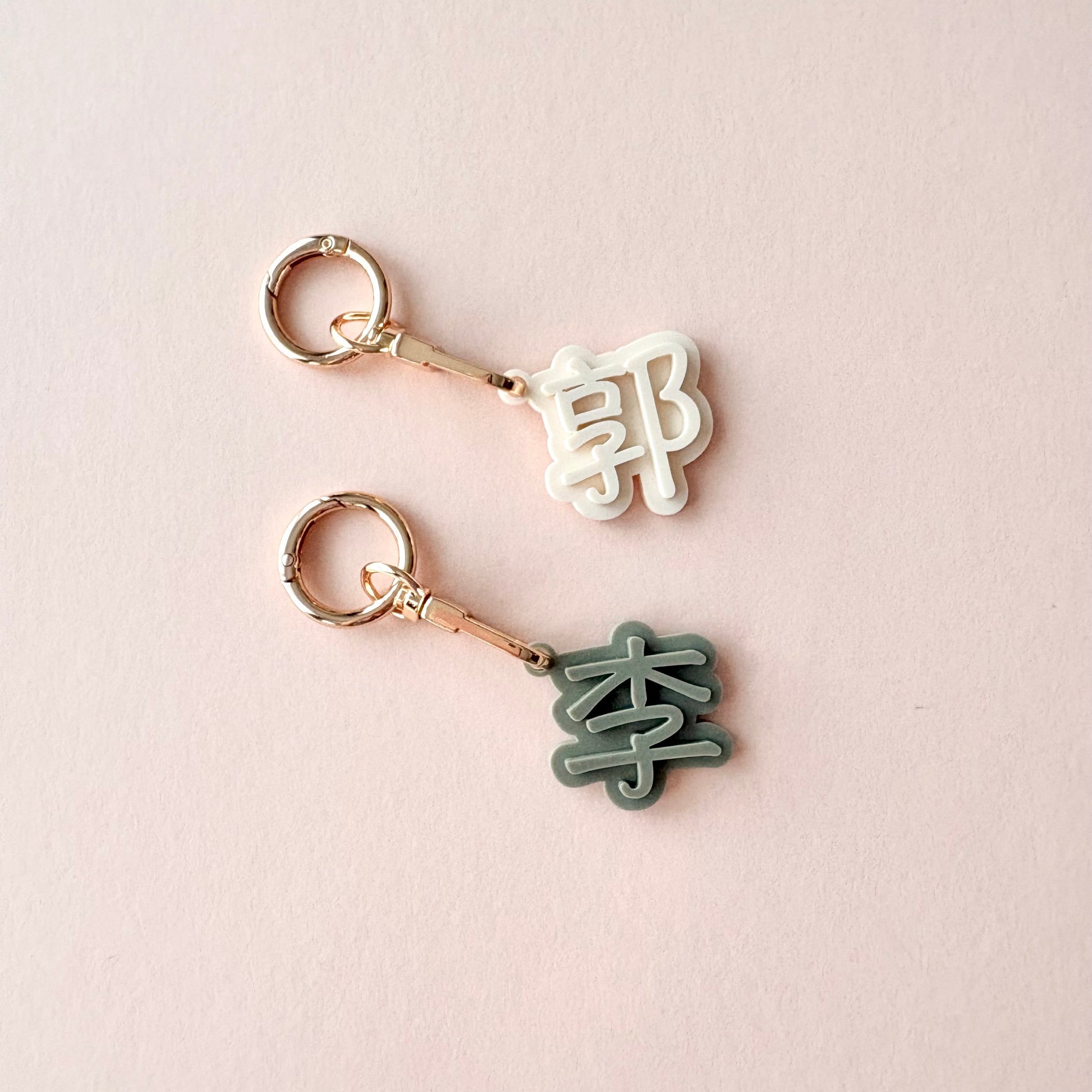 Two small metal keychains with Chinese character charms on a pale pink background. One charm is white and the other is dark green, both attached to rose gold keyrings.