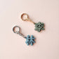 Two small keychain charms shaped like Chinese characters, one in light blue and one in muted green, attached to metal key rings on a pale pink background.