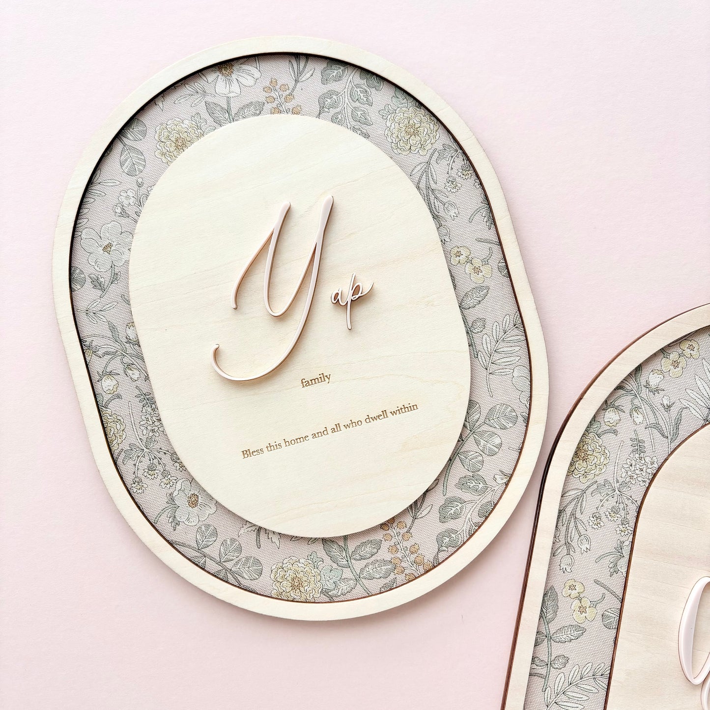 A close-up of a wooden personalized wall plaque or frame with a light natural wood oval center, a floral-patterned backing, and a small rose-gold monogram letter 'Y' with the word 'family' and a tiny heart. Part of a matching second plaque is visible on the right against a soft pink background.