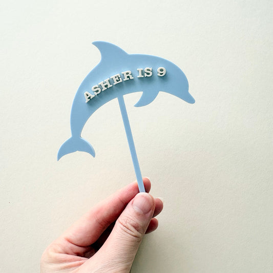 A hand holding a light blue dolphin-shaped cake topper with the text 'A.SHEER IS 9' in raised white letters, against a plain light beige background.