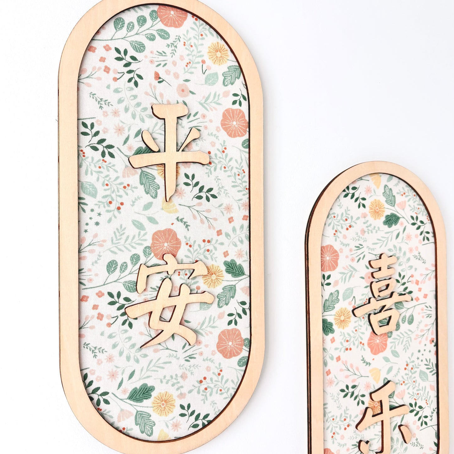 Two vertical oval wall decorations with light wood frames, featuring a delicate floral pattern in soft pink, orange, and green on a white background, with gold Chinese characters.
