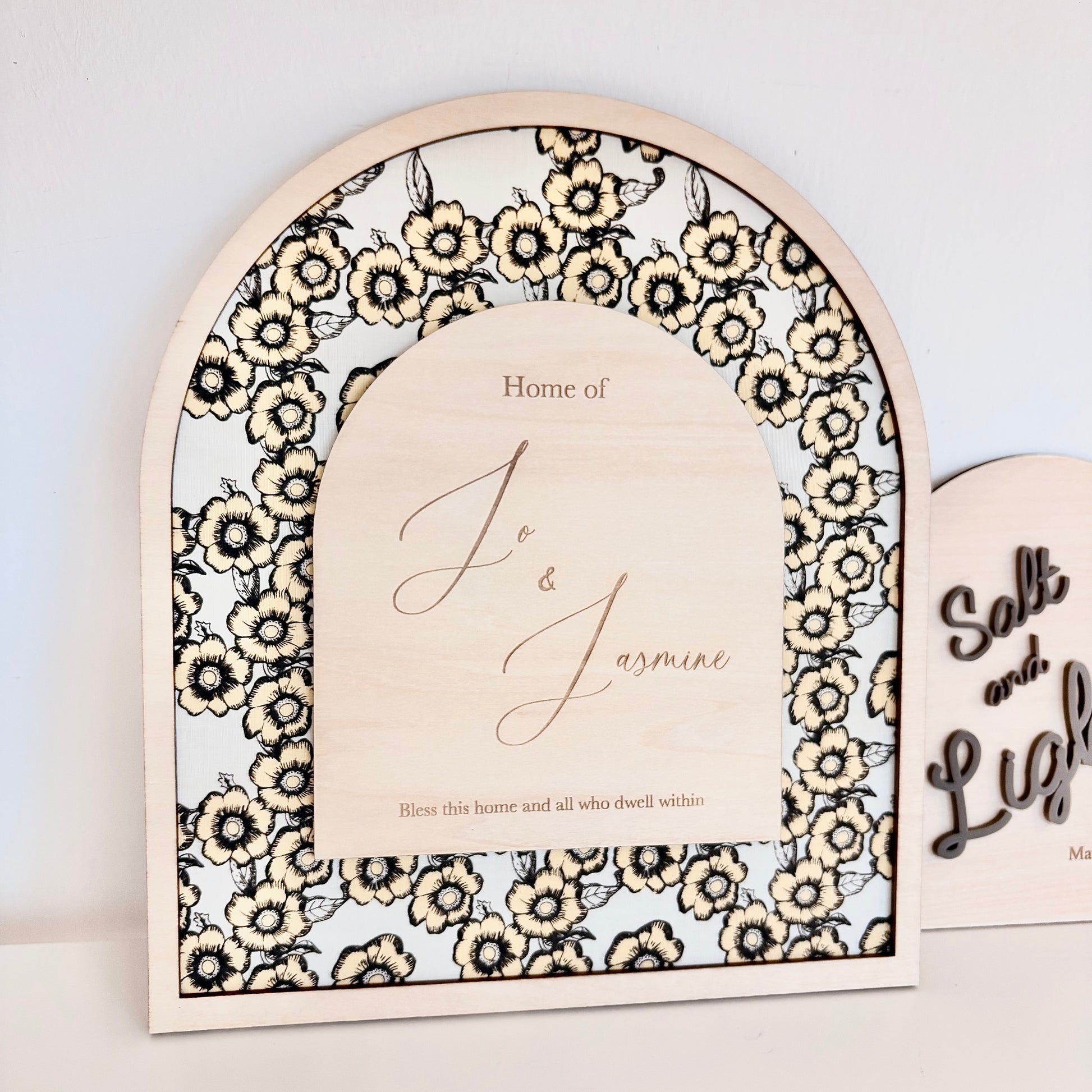 An arched wooden decorative sign with a black and cream floral border and a central light wood panel reading 'Home of J & Jasmine', shown against a plain light wall with part of another sign visible at the right edge.