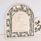 An arched wooden decorative sign with a black and cream floral border and a central light wood panel reading 'Home of J & Jasmine', shown against a plain light wall with part of another sign visible at the right edge.