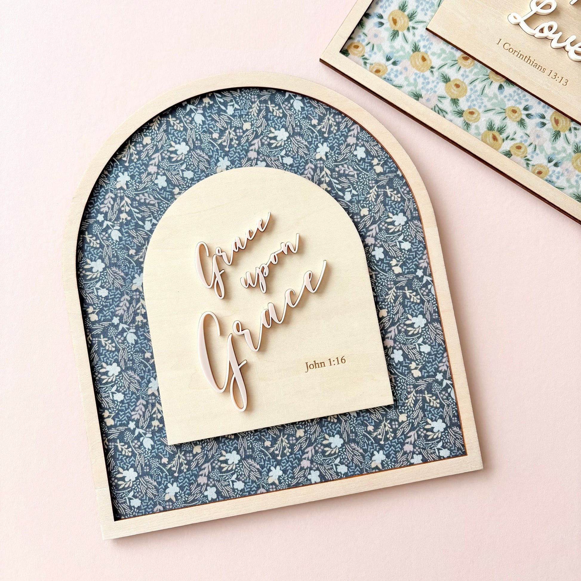 An arched wooden plaque with a blue floral patterned background and a light wood central panel reading 'Give me Grace' and 'John 1:16', styled on a pale pink surface with another matching floral plaque partially visible at the top right.