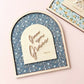 An arched wooden plaque with a blue floral patterned background and a light wood central panel reading 'Give me Grace' and 'John 1:16', styled on a pale pink surface with another matching floral plaque partially visible at the top right.