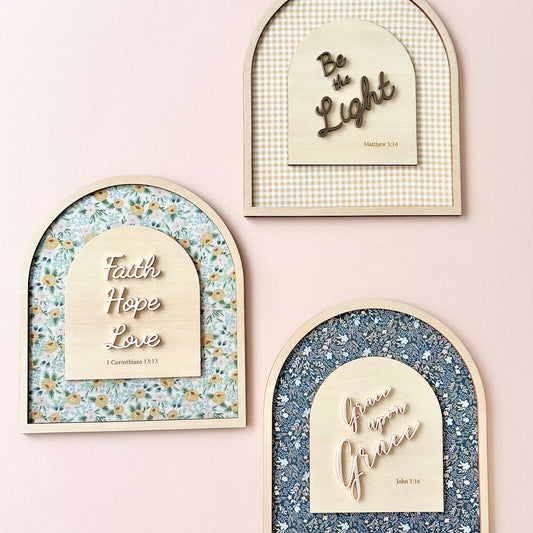 Three arched decorative wall art pieces mounted on a pale pink wall. Each piece has a light wood frame and a central cream panel with inspirational text: one says 'Be the Light,' one says 'Faith Hope Love,' and one says 'Grow upon Grace.
